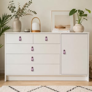 May include: A white wooden chest of drawers featuring six drawers and one cabinet door, each fitted with a small, light purple leather pull handle secured by a matching circular metal fastener, providing a functional and clean storage solution.