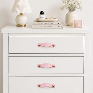 May include: A white three-drawer dresser featuring three light pink leather drawer pulls. Each pull is secured with two metallic gold-toned circular fasteners. The dresser top holds a pleated lamp, a small tray with a bottle, and two books.