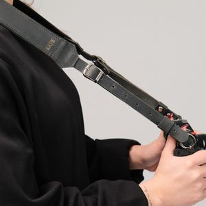May include: A black leather camera strap with the name "Kate" embossed on it. The strap is attached to a camera and is being held by a person.
