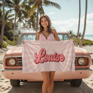 Könnte beinhalten: Ein rosa Strandtuch mit einem weißen Sonnenstrahlen-Design und dem Namen "Louise" in rosa Retro-Schrift.