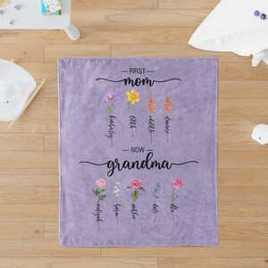 K&ouml;nnte beinhalten: Lilafarbene Fleece-Decke mit dem Text "First Mom" und einer Reihe von Blumen mit Namen darunter. Der Text "Now Grandma" befindet sich darunter mit einer weiteren Reihe von Blumen und Namen.
