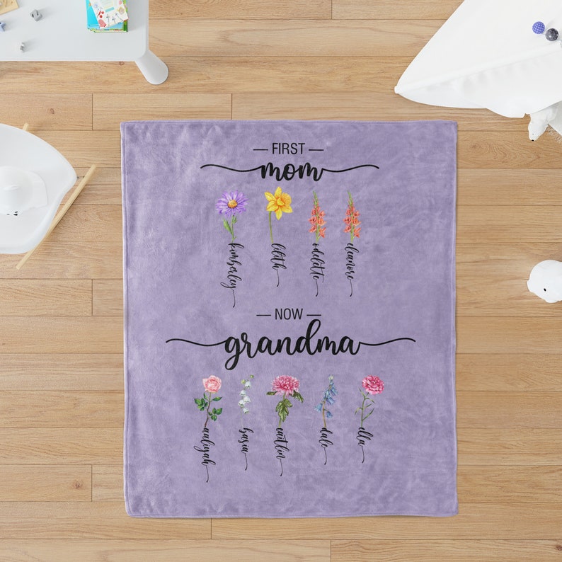 K&ouml;nnte beinhalten: Lilafarbene Fleece-Decke mit dem Text "First Mom" und einer Reihe von Blumen mit Namen darunter. Der Text "Now Grandma" befindet sich darunter mit einer weiteren Reihe von Blumen und Namen.