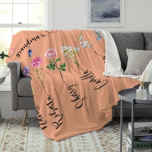 May include: Peach coloured fleece throw blanket with floral designs and the words "Grandma's", "Blessed", "Camille", and "We Love You" written in black cursive.