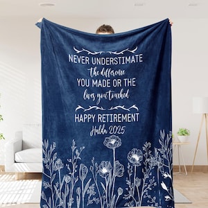 Puede incluir: Una manta azul marino con detalles florales blancos y el texto "NEVER UNDERESTIMATE the difference YOU MADE OR THE lives you touched HAPPY RETIREMENT Hulda 2025". La manta es sostenida por una persona.