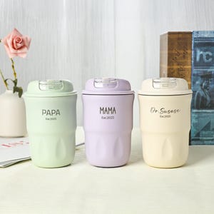 May include: Three insulated travel mugs in pastel colors: light green, lavender, and cream. Each mug has a matching lid and features text: "PAPA", "MAMA", and "Dr.Susase", with "Est.2025" below. The mugs are cylindrical with a ridged design.