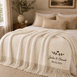 Könnte beinhalten: Eine cremefarbene Strickdecke mit Fransen, die eine dunkelbraune Stickerei mit dem Text Justin & Hannah 08.24.2026 sowie ein kleines Herz- und Blattzweigmotiv aufweist und über einem Bett mit Kissen ausgebreitet ist.