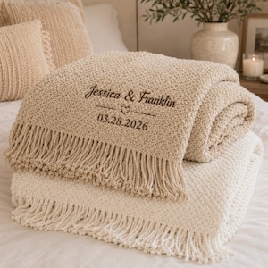 May include: A beige textured throw blanket with fringe edges, featuring dark brown embroidered text that reads Jessica & Franklin, a small heart icon, and the date 03.28.2026. The blanket is folded and layered over a cream-colored textured blanket.