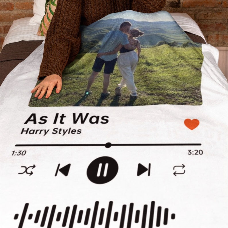 Op de afbeelding: Een wit deken met een foto van een stel in een veld, een Spotify-achtige muziekspelerinterface met het nummer "As It Was" van Harry Styles en een rood hartpictogram.
