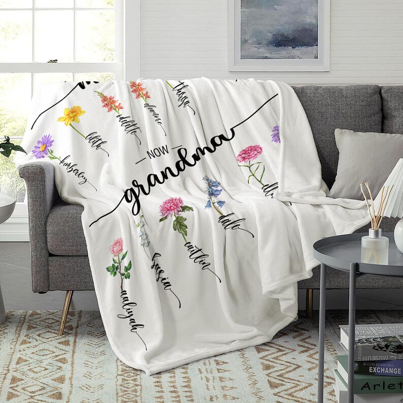 K&ouml;nnte beinhalten: Wei&szlig;e Fleece-Decke mit Blumenmuster und dem Text "Jetzt Oma" mit den Namen der Enkelkinder, die um die Blumen herum geschrieben sind.