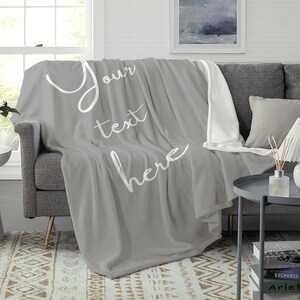 May include: A grey fleece throw blanket with white text that says "Your text here". The blanket is folded and draped over a grey sofa.