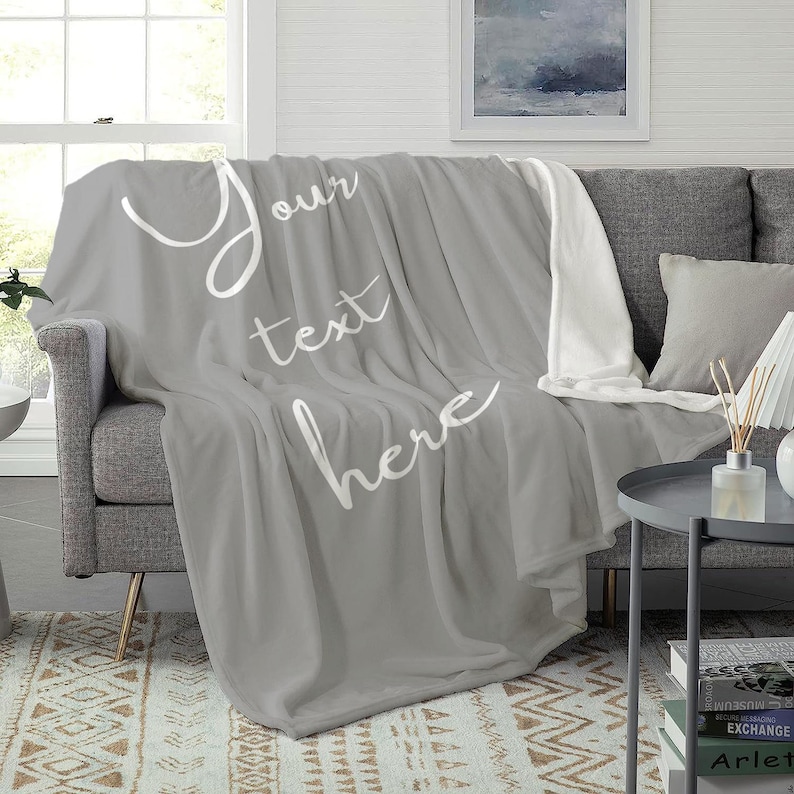 May include: A grey fleece throw blanket with white text that says "Your text here". The blanket is folded and draped over a grey sofa.