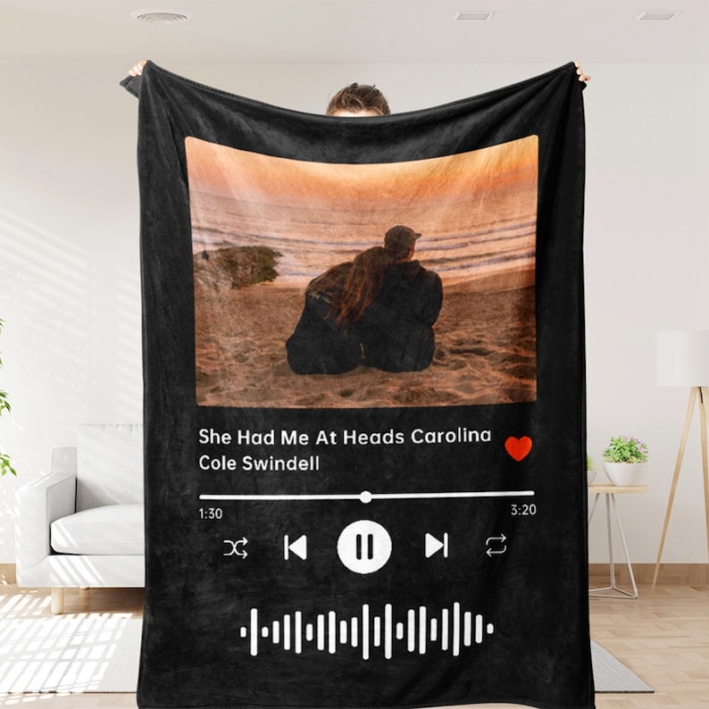 Op de afbeelding: Zwarte fleece deken met een foto van een stel dat op een strand bij zonsondergang zit. De deken heeft een Spotify-achtig muziekspelerontwerp met de titel van het nummer "She Had Me At Heads Carolina" van Cole Swindell, een rood hartpictogram en een afspeelbalk met tijdstempels.