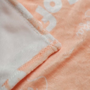 Op de afbeelding: Een perzikkleurig fleece deken met een witte rand en witte tekst. De deken is dubbelgevouwen, waardoor de zachte, pluche textuur zichtbaar is.