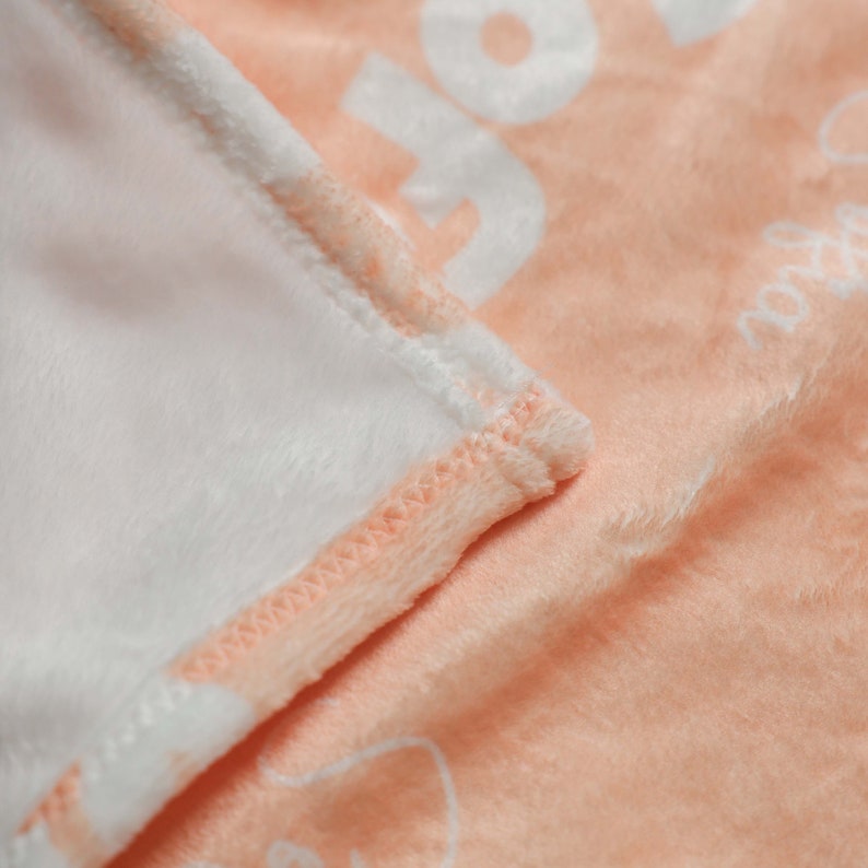 Op de afbeelding: Een perzikkleurig fleece deken met een witte rand en witte tekst. De deken is dubbelgevouwen, waardoor de zachte, pluche textuur zichtbaar is.