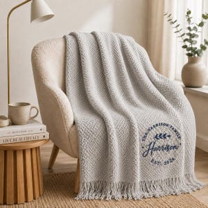 May include: A light grey textured knit throw blanket with fringe trim, featuring a circular navy blue embroidered design that reads The Harrison Family, Harrison, and Est. 2026, draped over a cream-coloured upholstered armchair.
