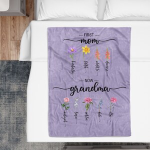 K&ouml;nnte beinhalten: Lilafarbene Fleece-Decke mit dem Text "First Mom" und "Now Grandma" mit Blumenillustrationen und den Namen der Kinder unter jeder Blume.