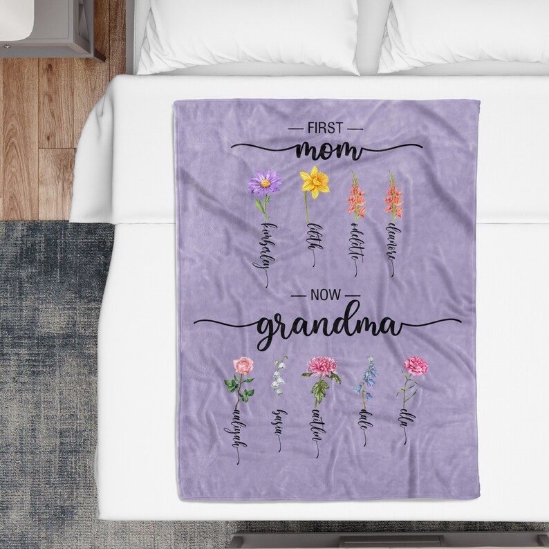 K&ouml;nnte beinhalten: Lilafarbene Fleece-Decke mit dem Text "First Mom" und "Now Grandma" mit Blumenillustrationen und den Namen der Kinder unter jeder Blume.