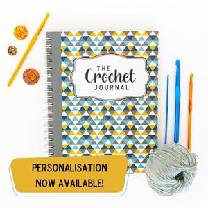 May include: A crochet journal with a geometric pattern in yellow, blue, and grey, with the title "The Crochet Journal". Crochet hooks, yarn, and decorative pins are arranged around the journal. A yellow banner states "PERSONALISATION NOW AVAILABLE!"