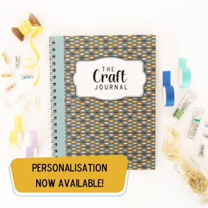 May include: A grey and blue patterned journal with a white label that says "The Craft Journal". The journal is surrounded by craft supplies, including yarn, tape, and paint tubes. The text "Personalisation Now Available!" is on a yellow banner.