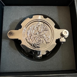 May include: A circular, metallic silver-toned gear-shaped object featuring an embossed dragon clutching a twenty-sided die with the number 20 visible. The object has a small mechanical hinge on the right side and is set within a black foam insert.