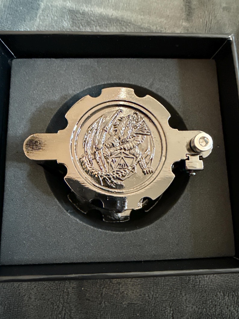May include: A circular, metallic silver-toned gear-shaped object featuring an embossed dragon clutching a twenty-sided die with the number 20 visible. The object has a small mechanical hinge on the right side and is set within a black foam insert.