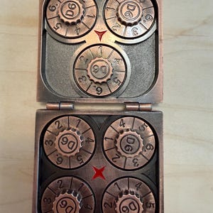 May include: A copper-colored metal case containing five circular rotating dials, each marked with the text D6 and numbers one through six. The top section features one dial with a red triangle indicator, while the bottom section holds four dials.