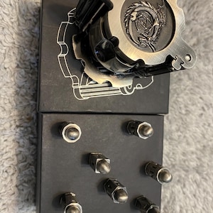 May include: A metallic, gear-shaped dice roller featuring an embossed dragon and a twenty-sided die design on its top surface. Below it, a black rectangular base holds seven small, silver-toned metal bullet-shaped game pieces arranged in a grid.