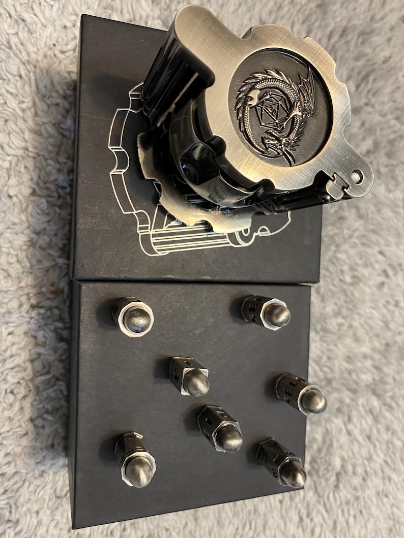 May include: A metallic, gear-shaped dice roller featuring an embossed dragon and a twenty-sided die design on its top surface. Below it, a black rectangular base holds seven small, silver-toned metal bullet-shaped game pieces arranged in a grid.