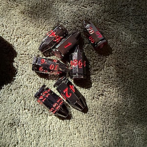 May include: A set of six bullet-shaped metal dice with a dark gunmetal finish and bright red engraved numbers. Each die features a pointed tip and a flat base, with various numerical values displayed in a clear, sans-serif font across the sides.