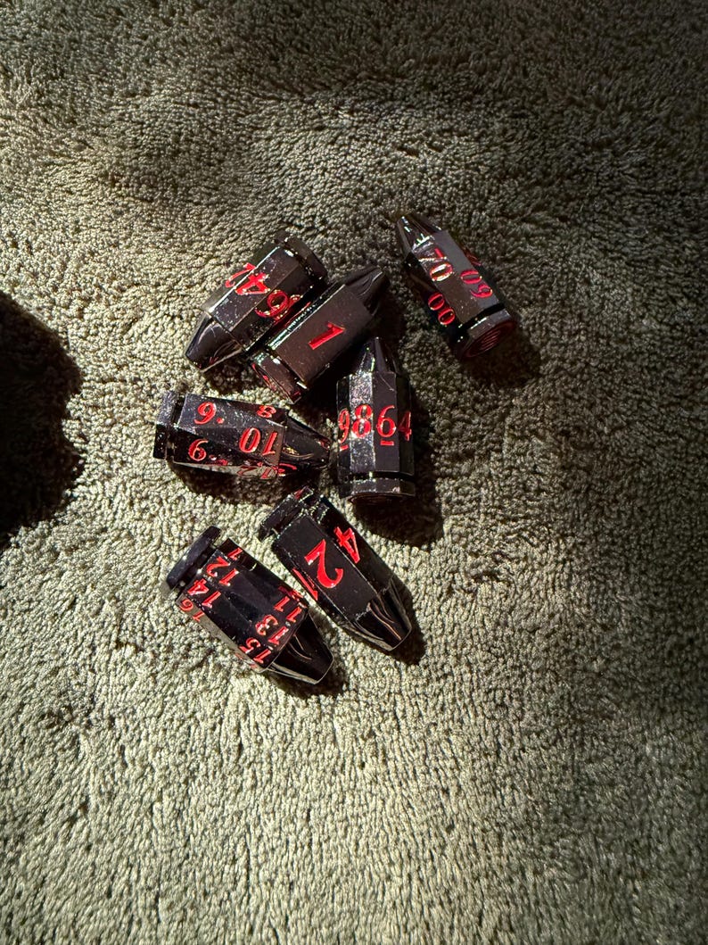 May include: A set of six bullet-shaped metal dice with a dark gunmetal finish and bright red engraved numbers. Each die features a pointed tip and a flat base, with various numerical values displayed in a clear, sans-serif font across the sides.