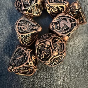 May include: A set of six black and copper metal dice with intricate skull and skeletal designs. Each die has a different number from 1 to 6.