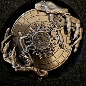 May include: A silver-colored, circular metal game piece with a sunburst design at the center. The piece features a clock-like outer ring with numbers and is flanked by stylized hands. The center has a D20 die and the text "D12 D6".