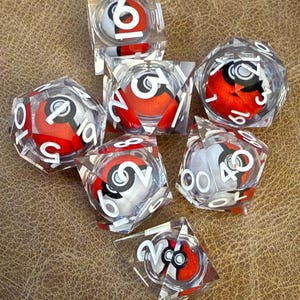 May include: A collection of clear, multi-sided dice with red and white spherical designs inside. Each die features white numbers and a black and white graphic. The dice are arranged on a textured brown surface.
