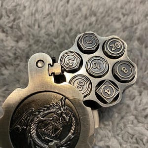 May include: A metallic, revolver-style dice holder with a hinged cover featuring an embossed dragon design. The open cylinder reveals six individual dice, each shaped like a hexagonal bolt, with numbers 4, 6, 8, 10, 12, and 20 clearly visible.