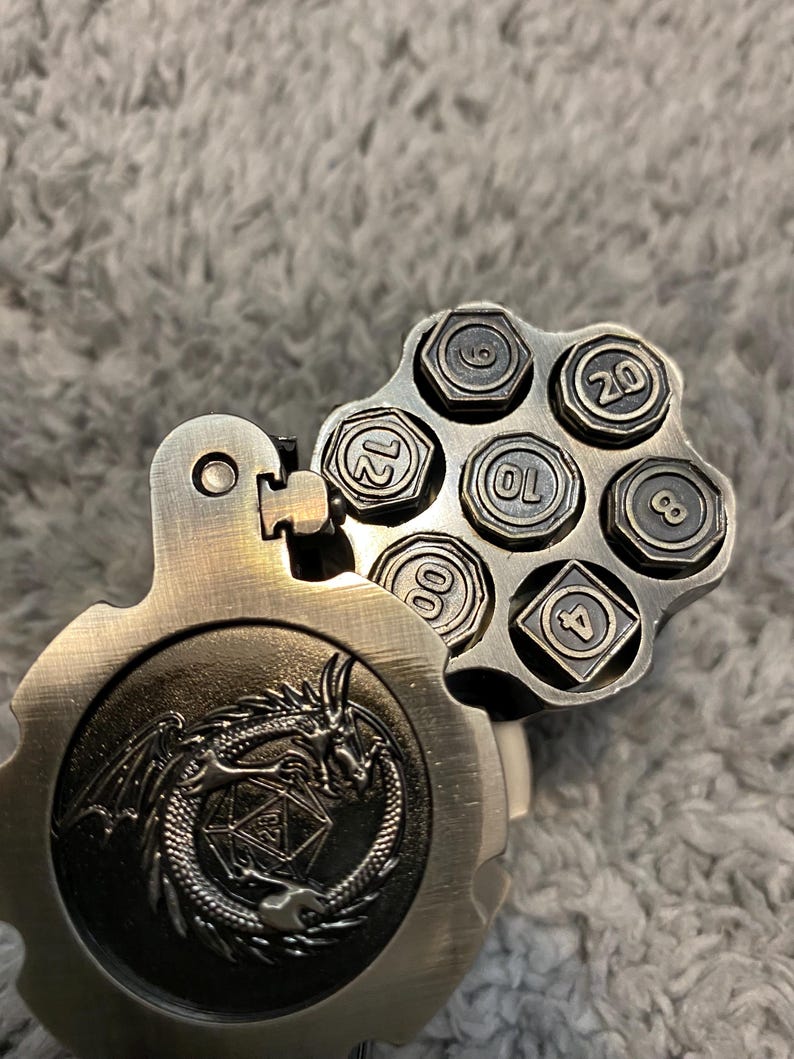 May include: A metallic, revolver-style dice holder with a hinged cover featuring an embossed dragon design. The open cylinder reveals six individual dice, each shaped like a hexagonal bolt, with numbers 4, 6, 8, 10, 12, and 20 clearly visible.