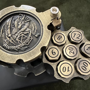 May include: A brass-colored metal dice holder shaped like a revolver cylinder, featuring a hinged lid embossed with a dragon and a twenty-sided die. The cylinder contains seven hexagonal dice inserts, each marked with numbers including 4, 6, 8, 10, 12, 20, and 00.