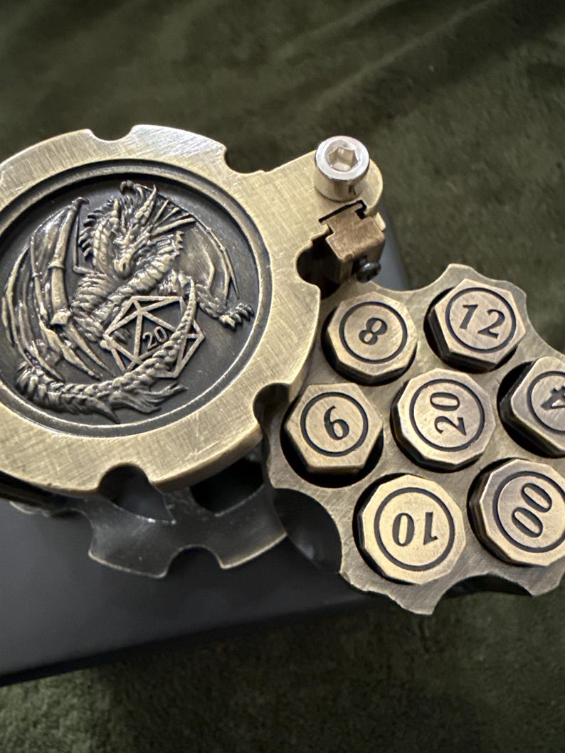 May include: A brass-colored metal dice holder shaped like a revolver cylinder, featuring a hinged lid embossed with a dragon and a twenty-sided die. The cylinder contains seven hexagonal dice inserts, each marked with numbers including 4, 6, 8, 10, 12, 20, and 00.