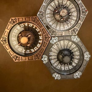 May include: Three hexagonal game wheels with numbers and decorative designs. The wheels are in bronze, silver, and gray tones. Each wheel has a central rotating dial and a glass cover, with numbers around the perimeter.