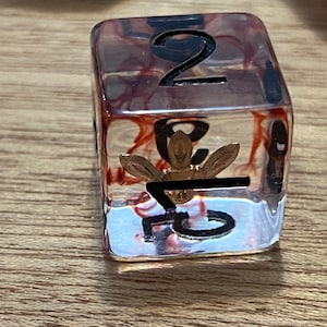 May include: A clear, red and gold dice with the number 2 on one side and the number 7 on another side.