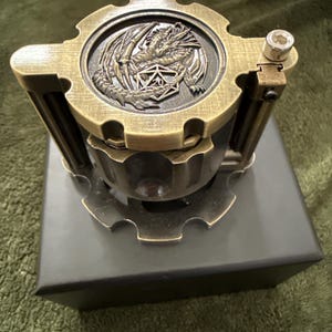 May include: A cylindrical metal dice container with a brushed brass finish. The lid features a raised relief of a dragon coiled around a twenty-sided die. The structure includes gear-shaped top and bottom plates with vertical support pillars.