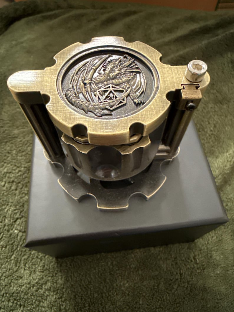 May include: A cylindrical metal dice container with a brushed brass finish. The lid features a raised relief of a dragon coiled around a twenty-sided die. The structure includes gear-shaped top and bottom plates with vertical support pillars.