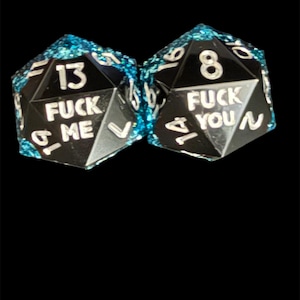 May include: Two black, multi-sided dice with white numbers and text. One die reads "13 FUCK ME" and the other reads "8 FUCK YOU". The edges are trimmed with a sparkling, light blue material.