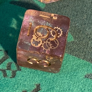 Puede incluir: Un dado transparente, iridiscente morado y rosa con engranajes dorados y el número 6 en la cara.