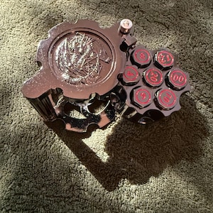 May include: A metallic silver-toned dice storage case shaped like a revolver cylinder with a gear-like lid featuring an embossed dragon design. The cylinder holds seven individual dice, each displaying red numbers including 4, 6, 8, 10, 12, 20, and 00.