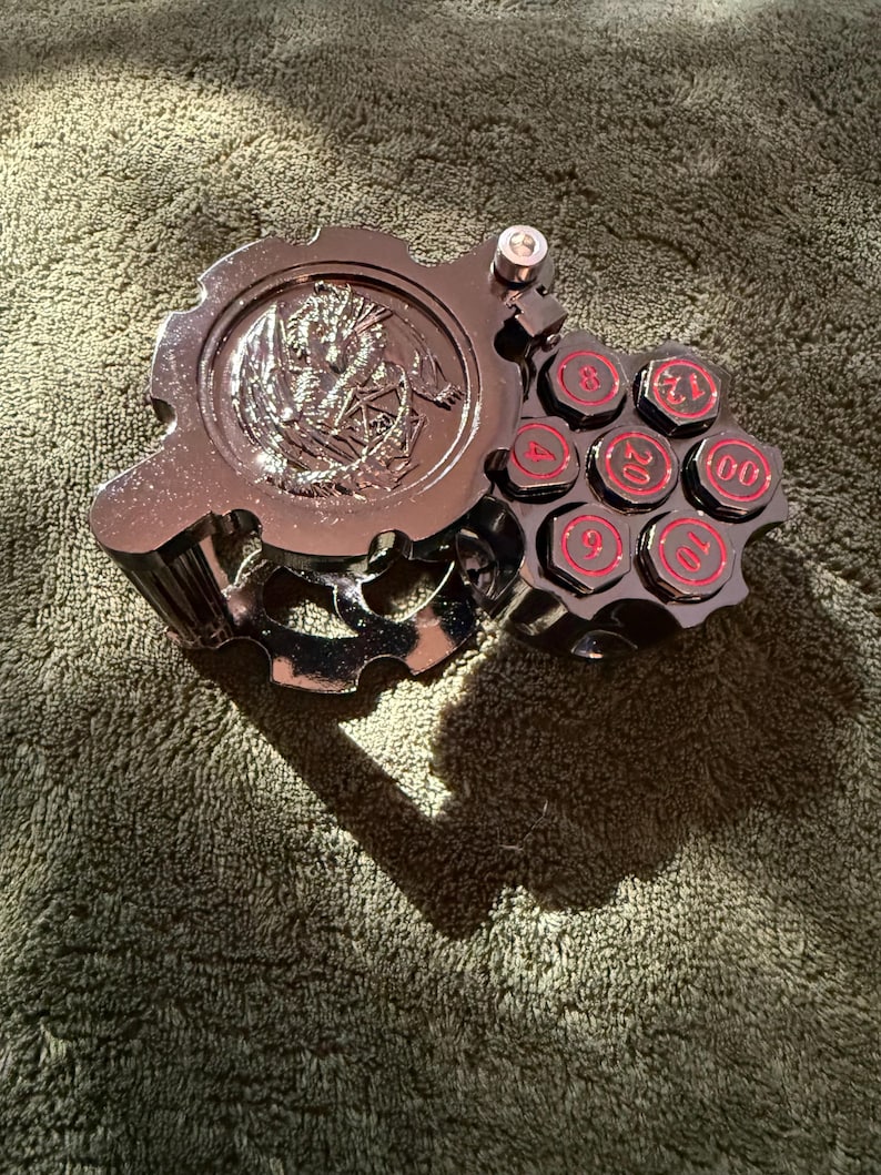 May include: A metallic silver-toned dice storage case shaped like a revolver cylinder with a gear-like lid featuring an embossed dragon design. The cylinder holds seven individual dice, each displaying red numbers including 4, 6, 8, 10, 12, 20, and 00.