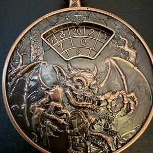 May include: A bronze-coloured, circular decorative piece with a devilish gargoyle design. The piece features a dial with numbers and the text "20 10 D8" at the top. The gargoyle has bat wings and is holding a chest.