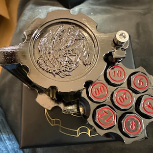 May include: A black metallic dice storage container shaped like a revolver cylinder with a dragon emblem on the lid. The cylinder features six circular slots containing dice faces marked with red numbers 00, 6, 4, 8, 12, and 20, plus a metal bolt.