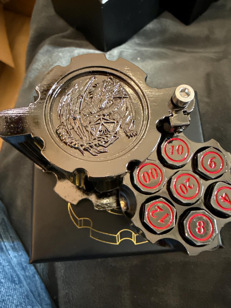 May include: A black metallic dice storage container shaped like a revolver cylinder with a dragon emblem on the lid. The cylinder features six circular slots containing dice faces marked with red numbers 00, 6, 4, 8, 12, and 20, plus a metal bolt.