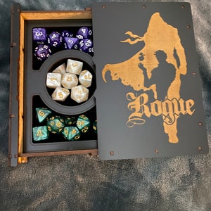 May include: A wooden box with a hinged lid, the lid is open and shows a silhouette of a person with the word "Rogue" underneath. The box contains a set of dice, including a set of purple dice, a set of green dice, and a set of white dice.