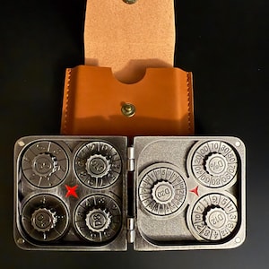 May include: A metal dice set with four dice on each side, each die with different numbers. The set is in a brown leather case with a snap closure. The dice are silver-colored and the case is brown.